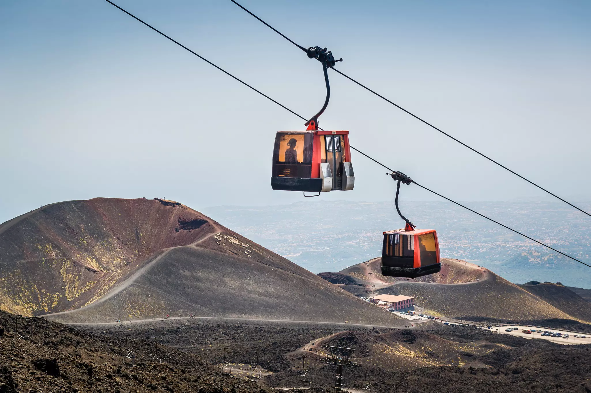 Two gondolas on a cable car pass one another. The slopes and craters of a volcano are visible below them.