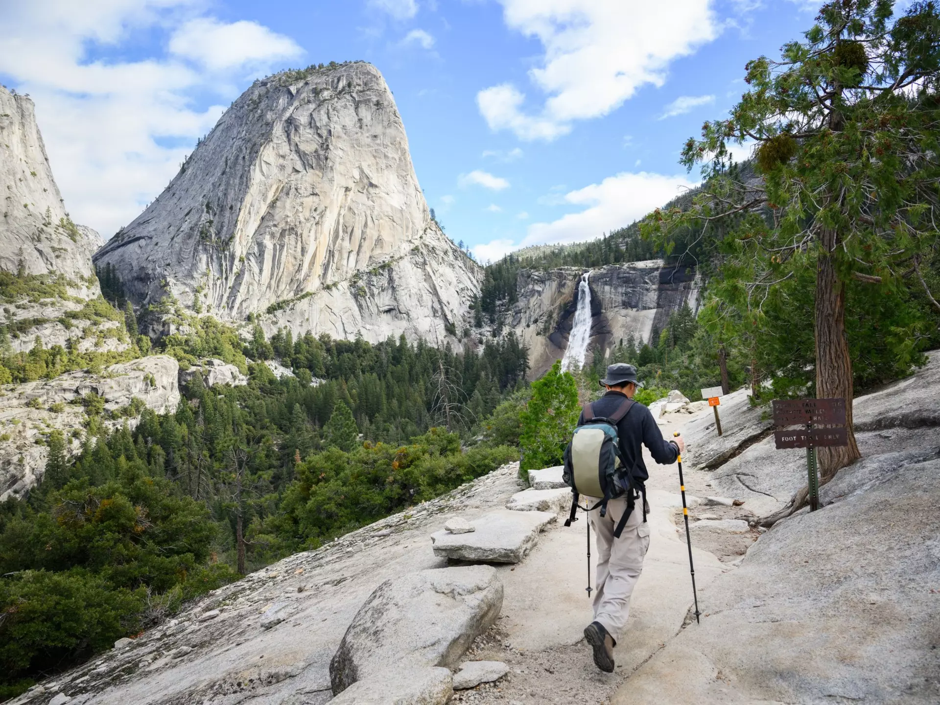 A hiker with poles is seen from behind walking on a rocky stretch of trail in a forest toward a waterfall.