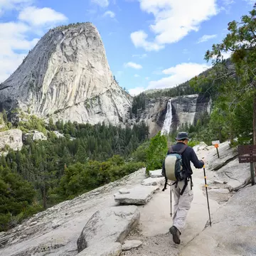 A hiker with poles is seen from behind walking on a rocky stretch of trail in a forest toward a waterfall.