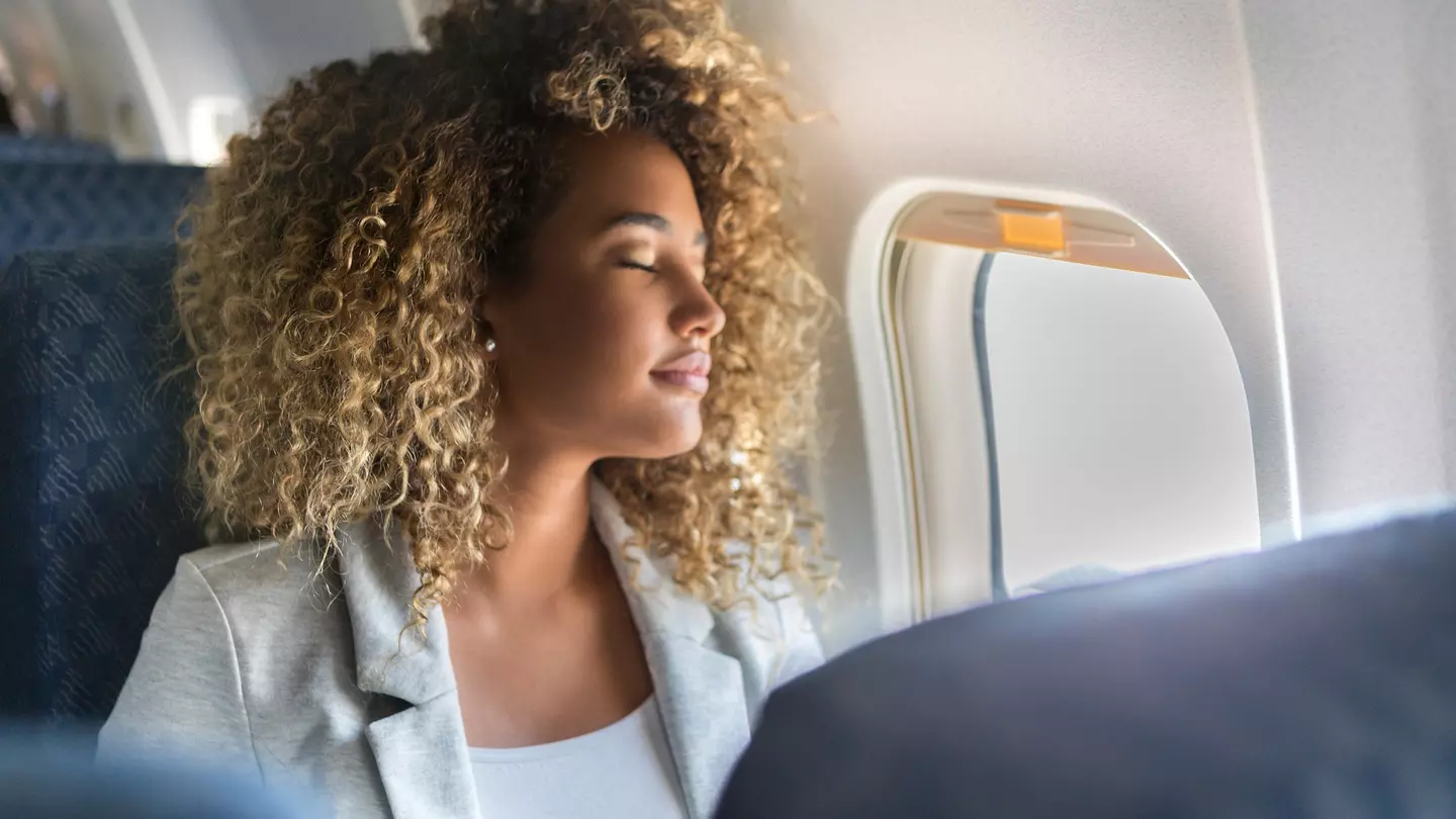 A young woman sits at the window seat of a commercial airliner and leans back with her eyes closed. SDI Productions / Getty Images