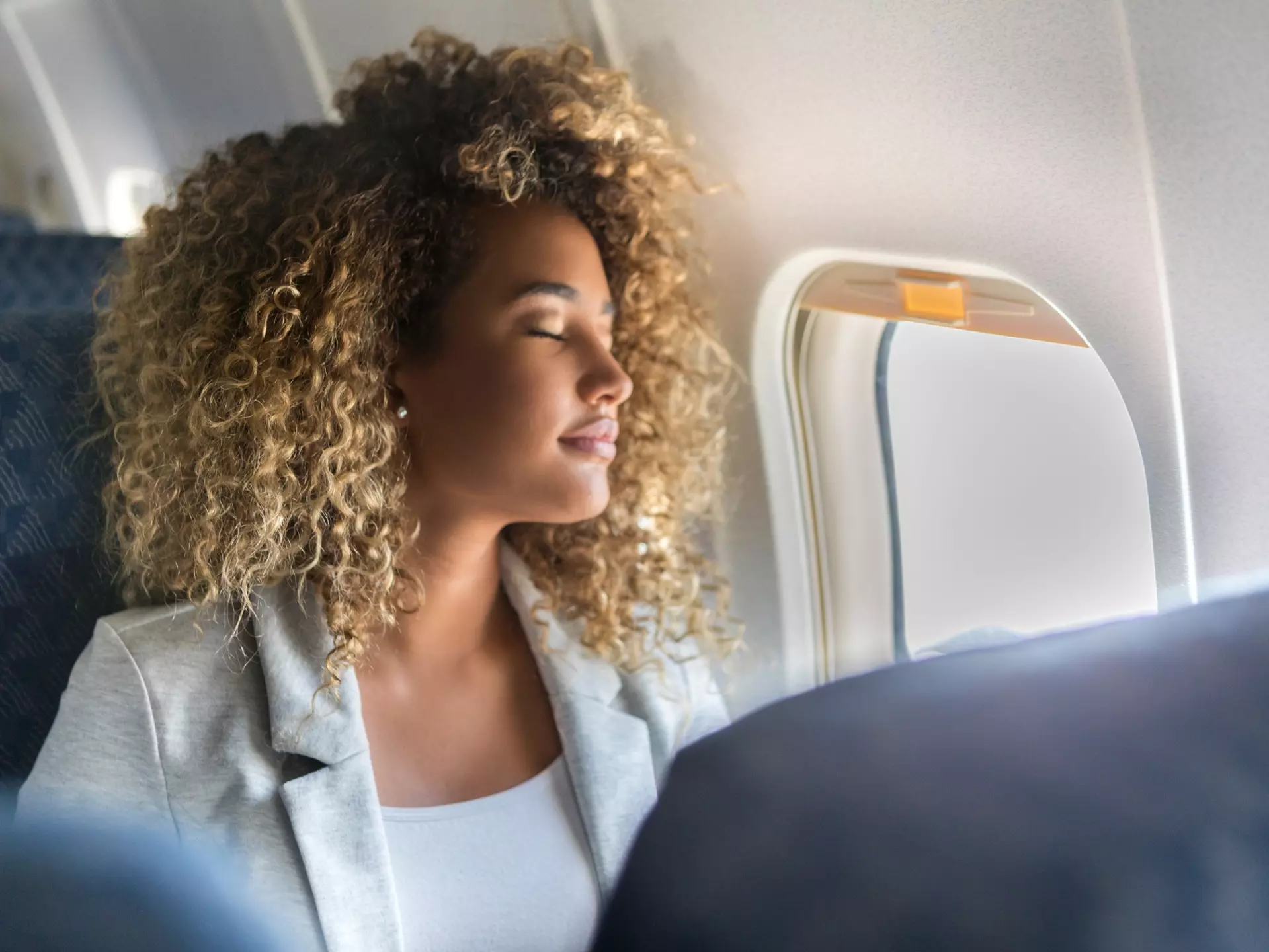 A young woman sits at the window seat of a commercial airliner and leans back with her eyes closed. SDI Productions / Getty Images