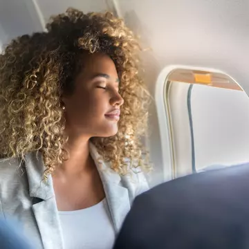 A young woman sits at the window seat of a commercial airliner and leans back with her eyes closed. SDI Productions / Getty Images