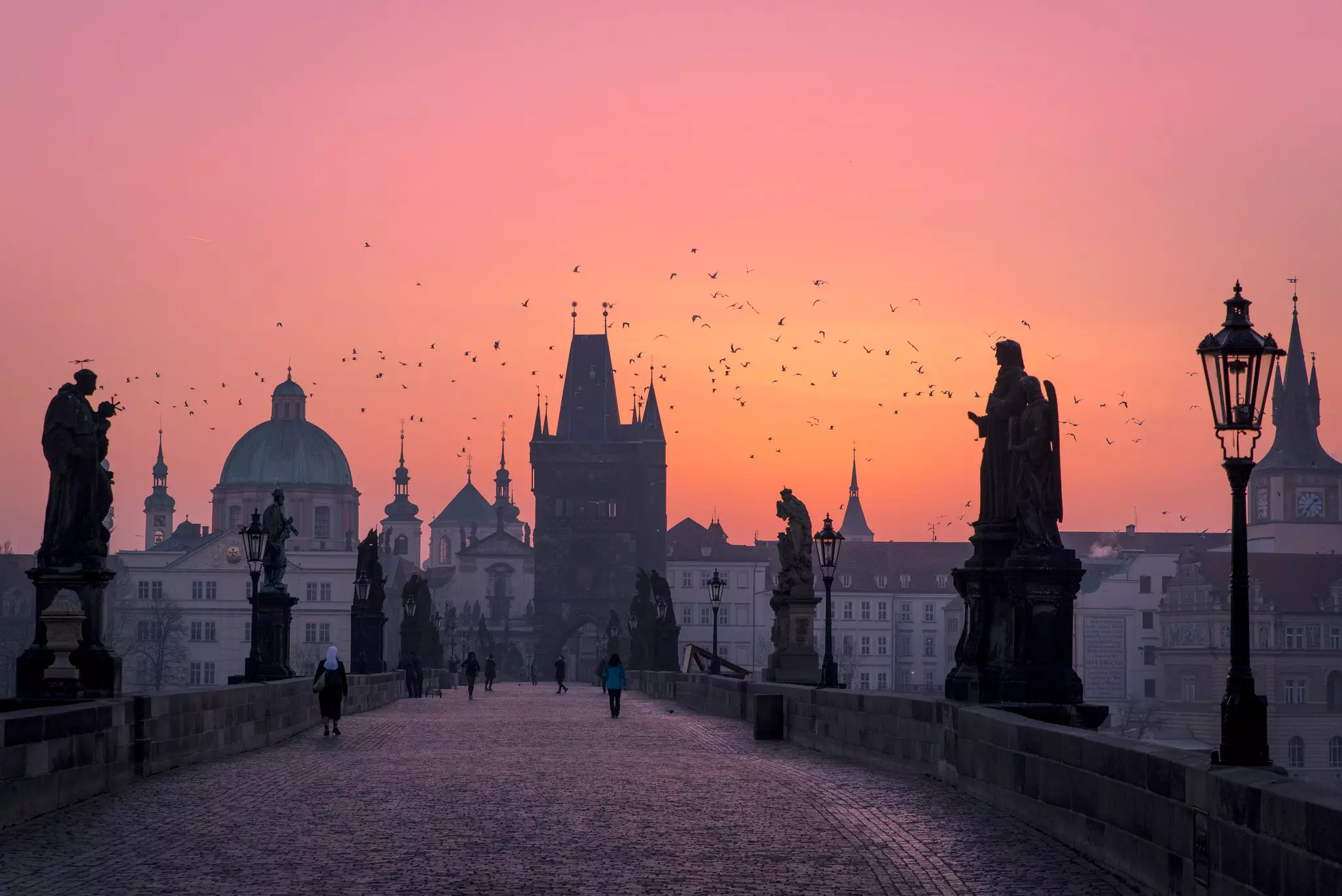 A pink, yellow and orange sky at sunrise over an old city, with people walking on a stone bridge lined with statues