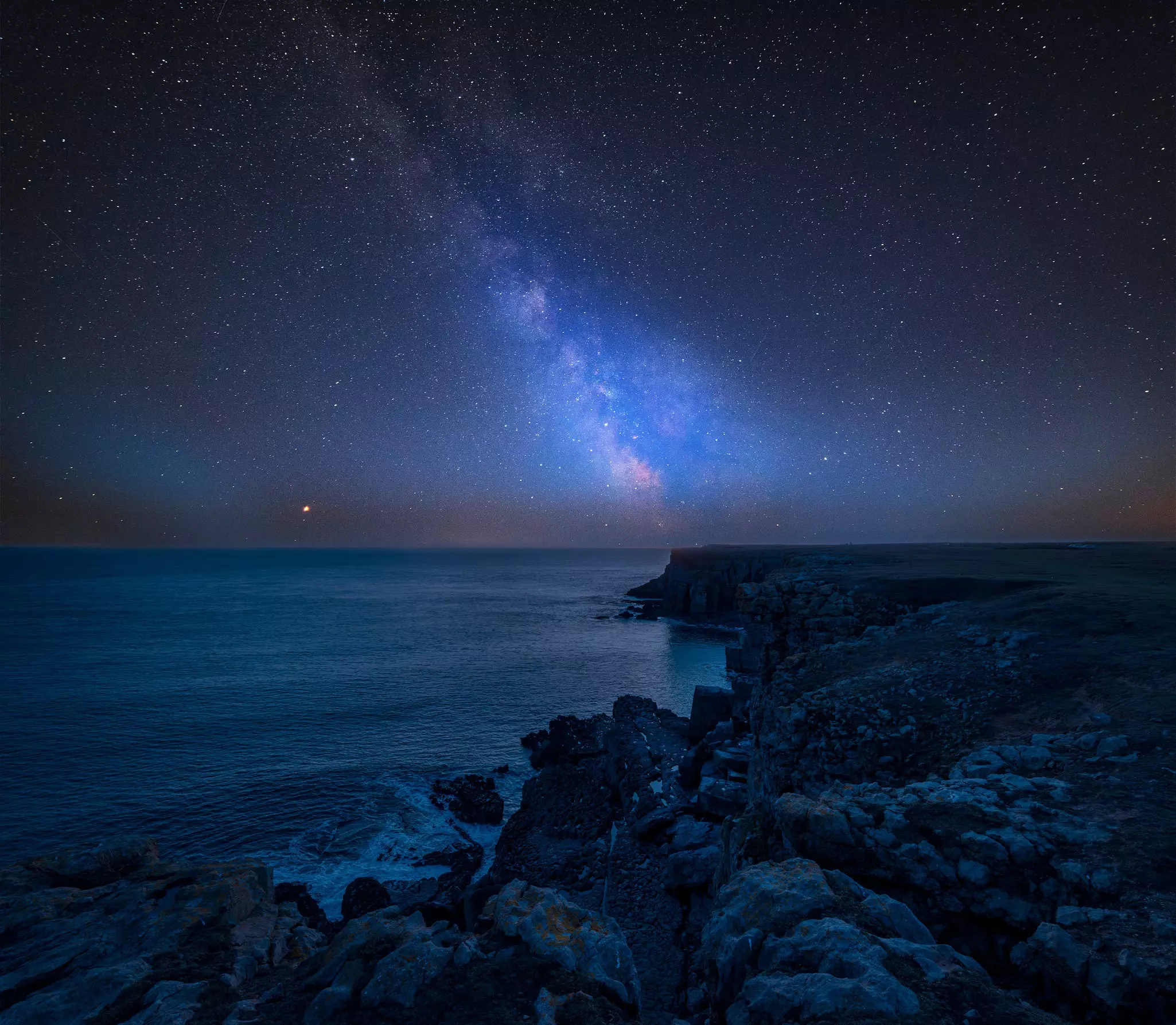 Thge Milky Way in the night sky over a rocky section of the coast. The water reflects the light.