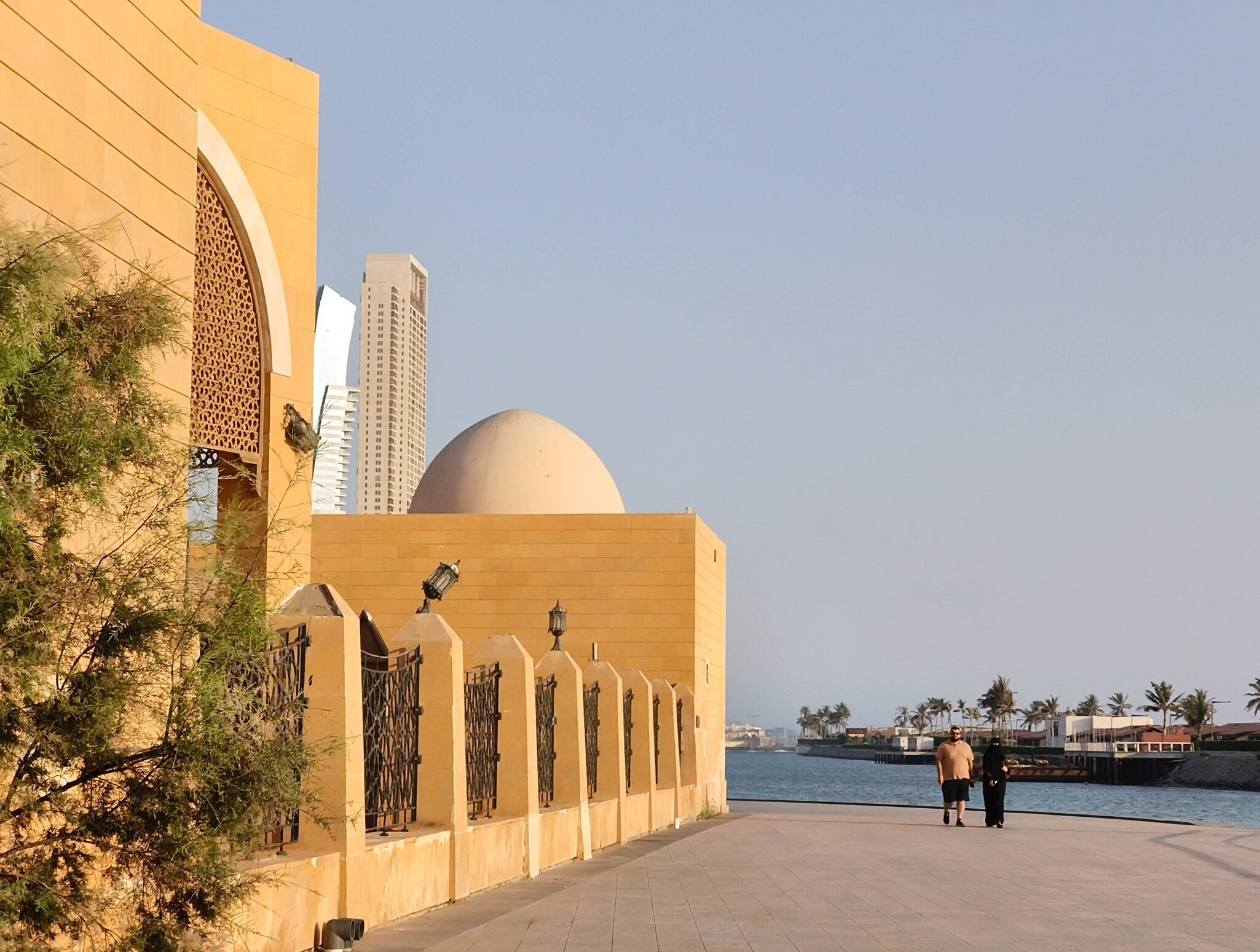 A couple walks the Jeddah Corniche past a sandstone mosque with a domed roof, with towers and the Red Sea behind