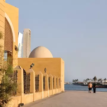 A couple walks the Jeddah Corniche past a sandstone mosque with a domed roof, with towers and the Red Sea behind