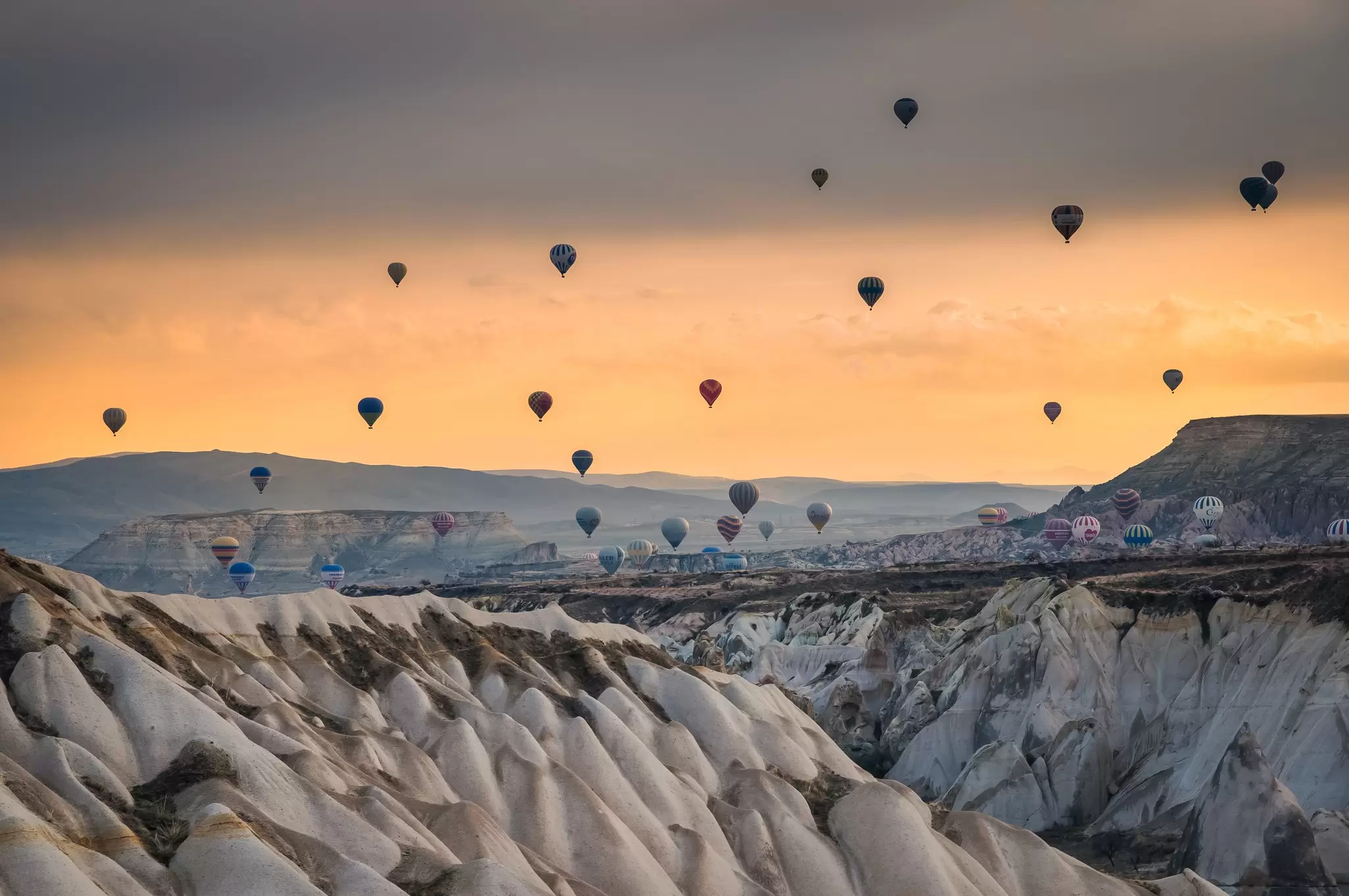 Dozens of hot air balloons drifting over eroded rock valleys at sunrise, evoking Cappadocia