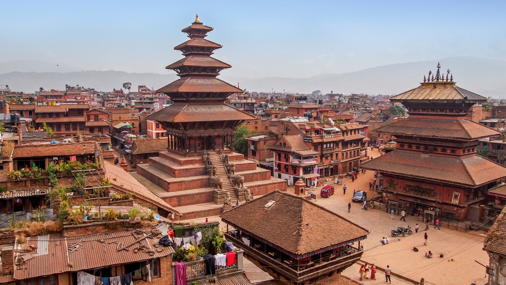 Tall five-roofed brick pagoda on a stepped plinth beside a wide temple and a square with people and brick houses, Bhaktapur.