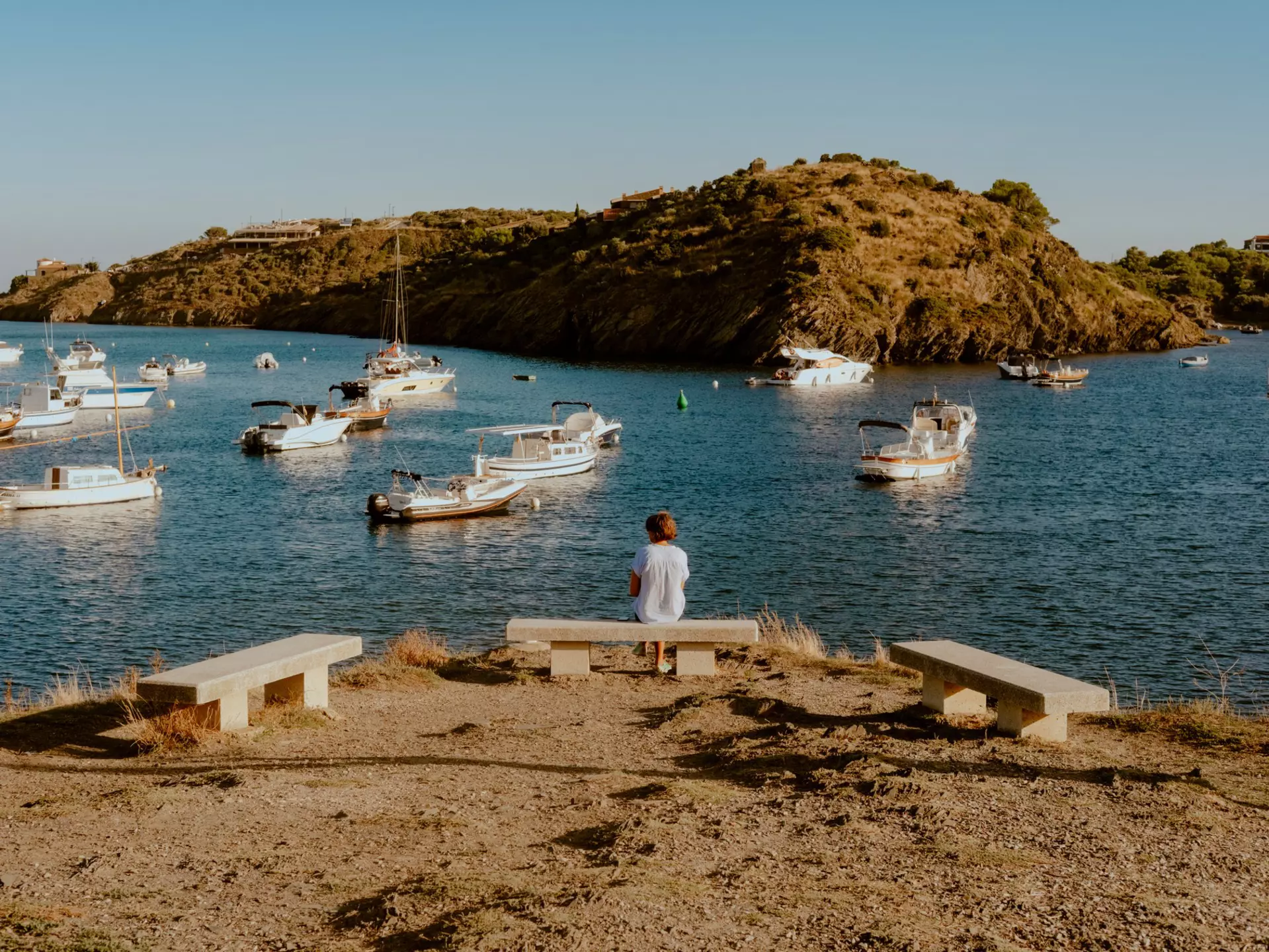 A person sits on a bench by a harbor with boats.