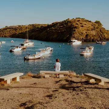 A person sits on a bench by a harbor with boats.