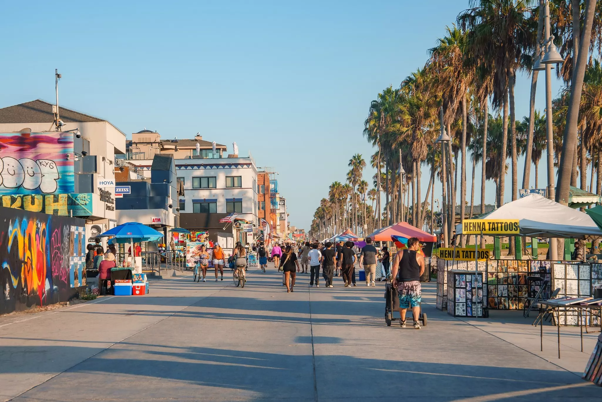 People walk and cycle along the vibrant Venice Beach Boardwalk in Los Angeles. Palm trees line the path, with colorful graffiti art on nearby walls.
