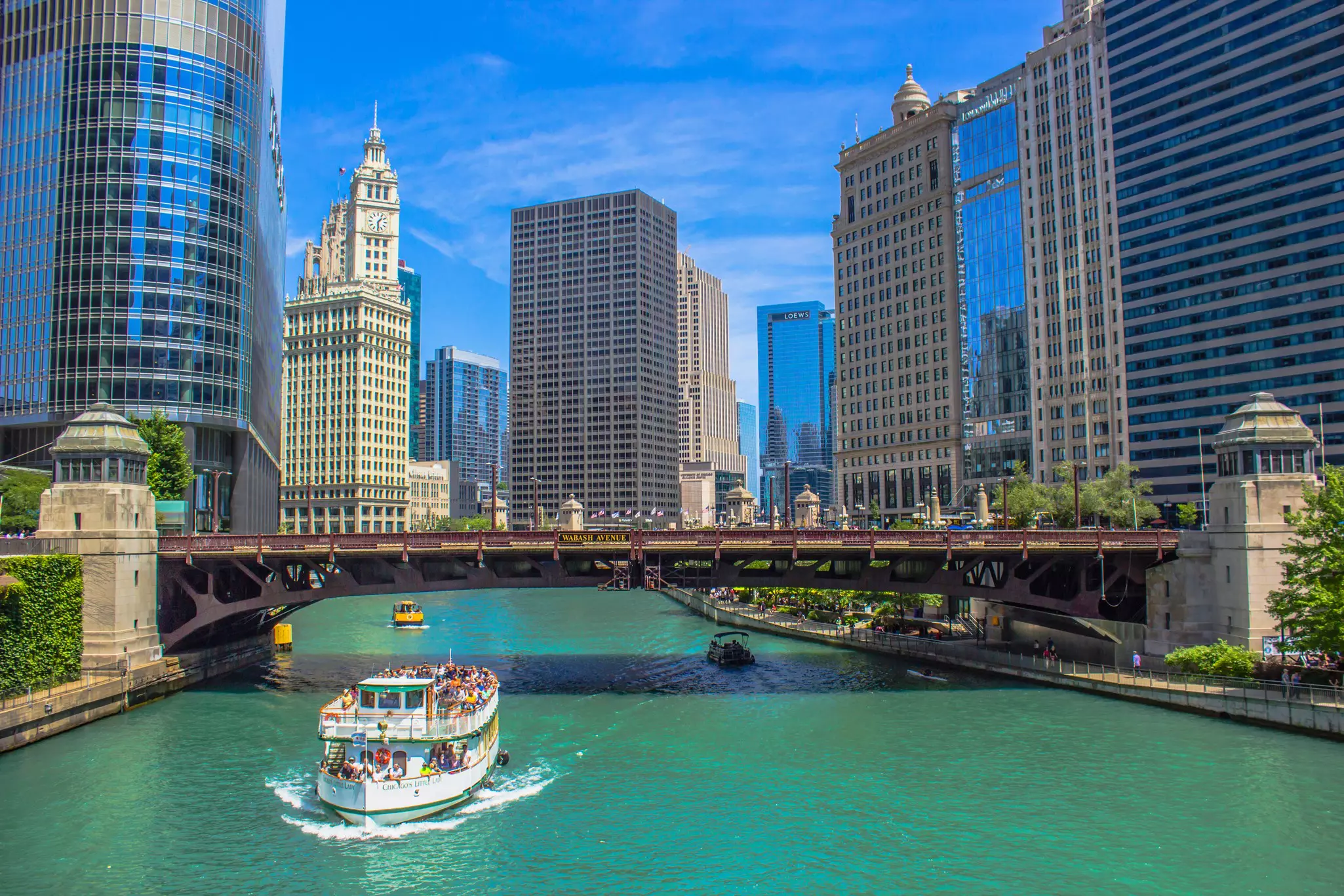 Boats loaded with tourists cruise along a river that flows between tall skyscrapers in a high-rise city