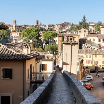Stone-walled elevated footpath running between ochre houses toward a hilltop town with bell towers in Perugia, Umbria.