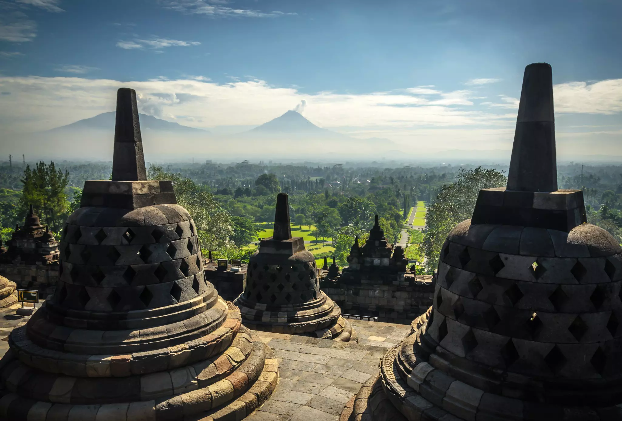 Bell-shaped stupa monuments on the upper level of a Buddhist temple stand above a green plain with distant peaks in Indonesia.