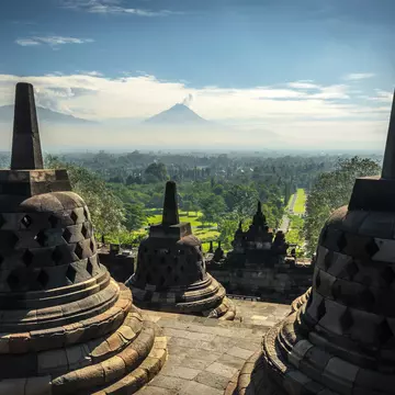 Bell-shaped stupa monuments on the upper level of a Buddhist temple stand above a green plain with distant peaks in Indonesia.