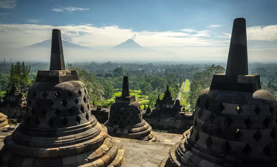 Bell-shaped stupa monuments on the upper level of a Buddhist temple stand above a green plain with distant peaks in Indonesia.