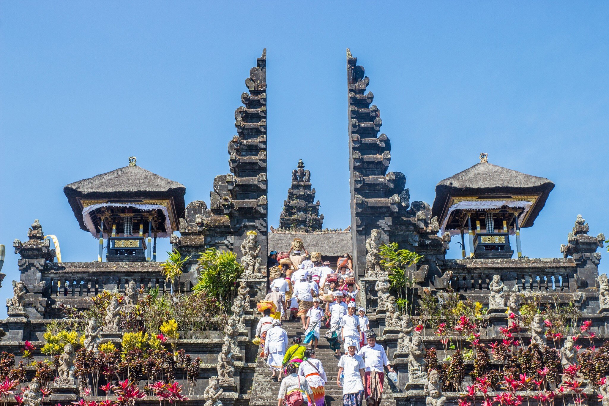 People in white clothing climbing temple steps through a tall carved stone split gateway, with pavilions either side.