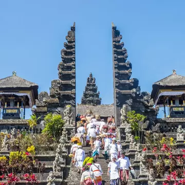 People in white clothing climbing temple steps through a tall carved stone split gateway, with pavilions either side.