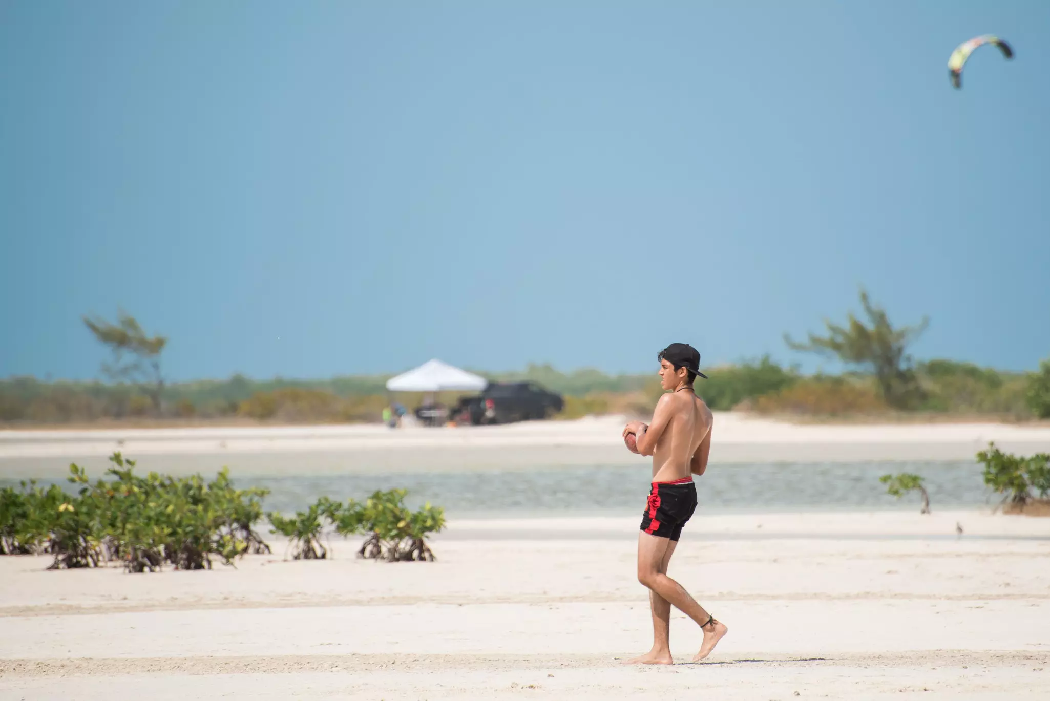 A young man in a swimsuit and baseball cap tosses a football on a deserted beach. A lagoon and vegetation are visible in the distance.