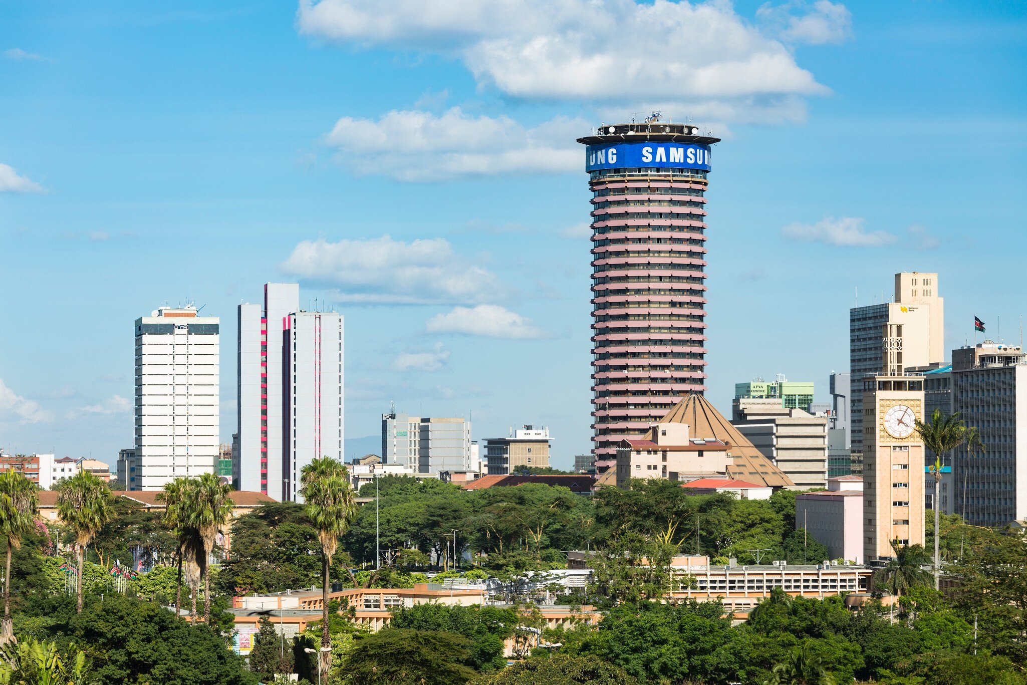 Tall cylindrical tower with banded balconies and a saucer-shaped crown above a green city of offices and palms.