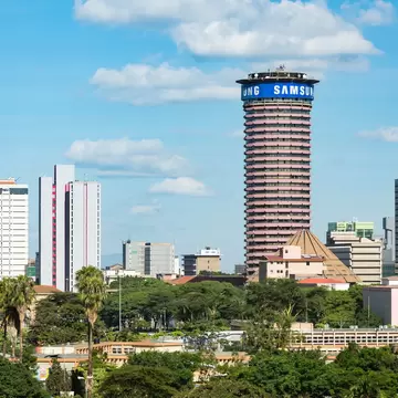 A tall cylindrical tower with banded balconies and a saucer-shaped crown standing over a green city of office blocks and palms.