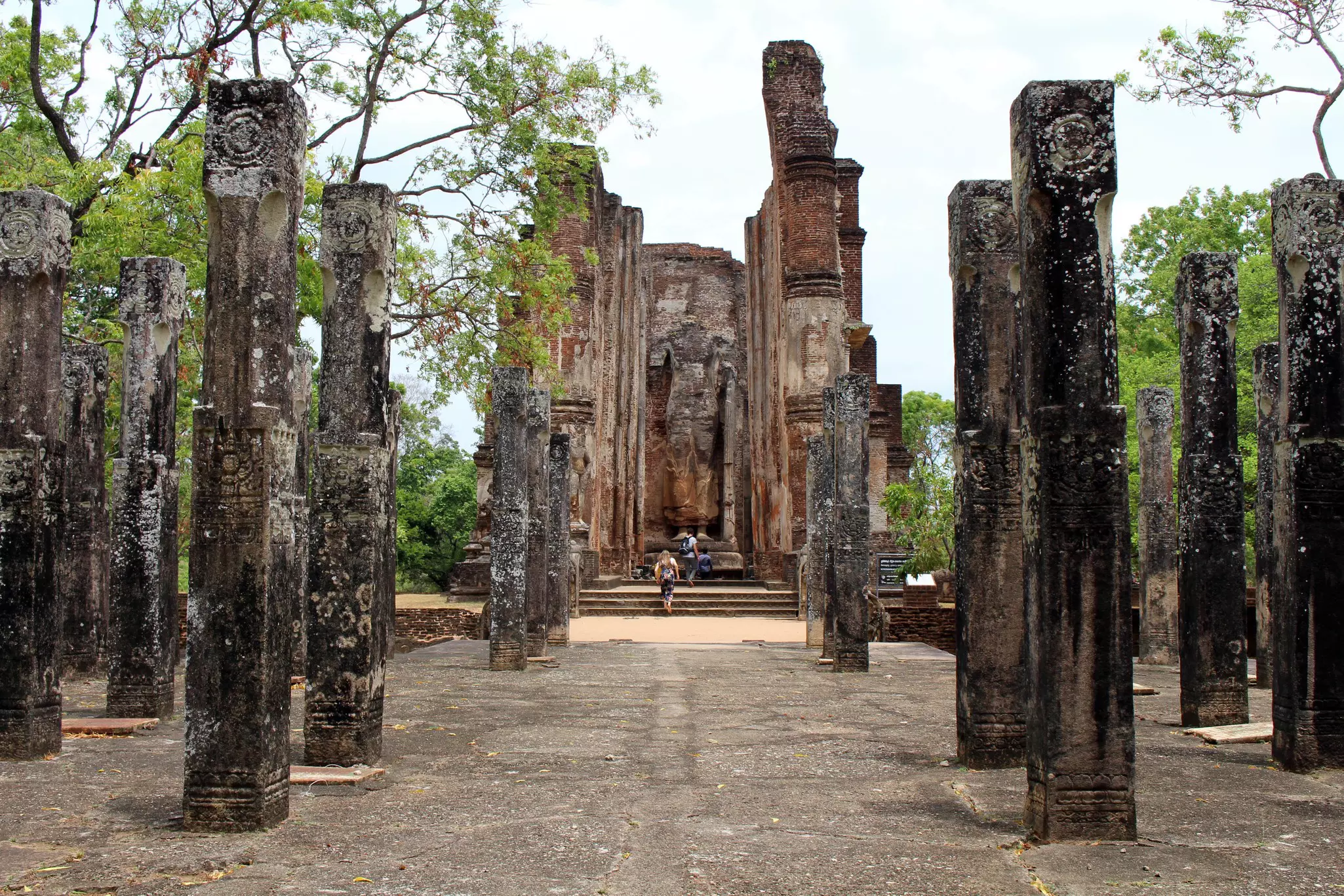 Two people walk between talk stone pillars with carvings towards the ruins of an ancient structure