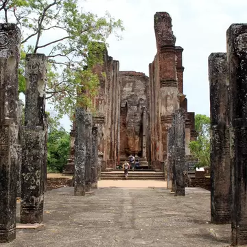 Two people walk between talk stone pillars with carvings towards the ruins of an ancient structure