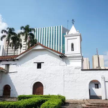 White colonial church with a bell tower and tiled roofs behind a clipped hedge, modern high-rises rising behind it.