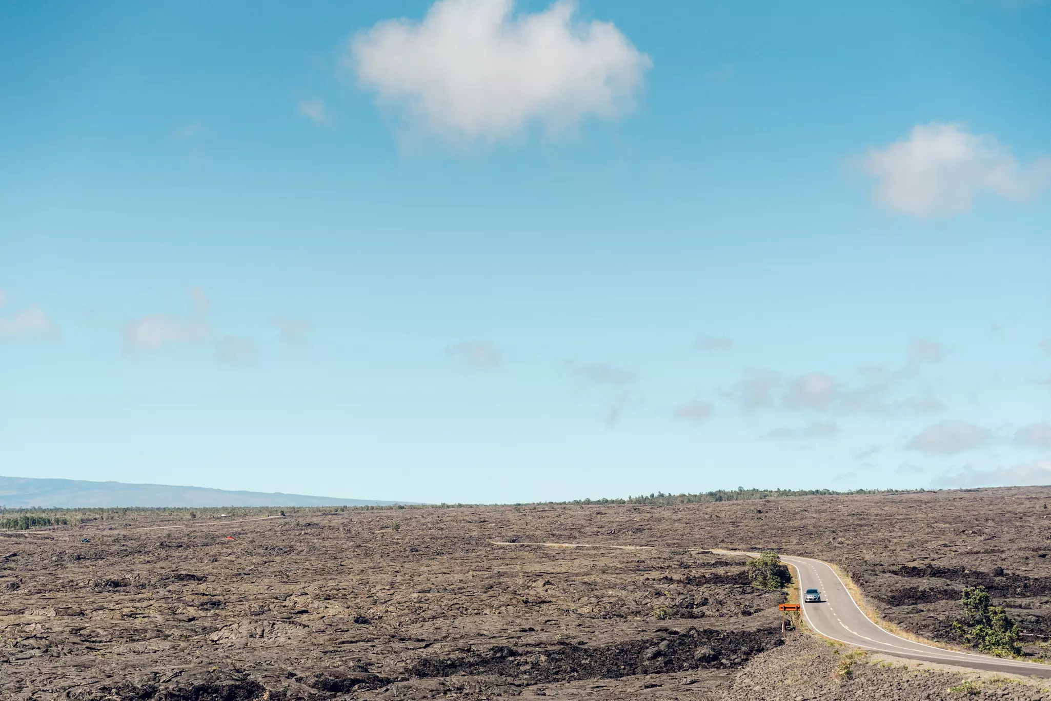 A car travels on a two-lane road through a barren volcanic landscape on a sunny day.
