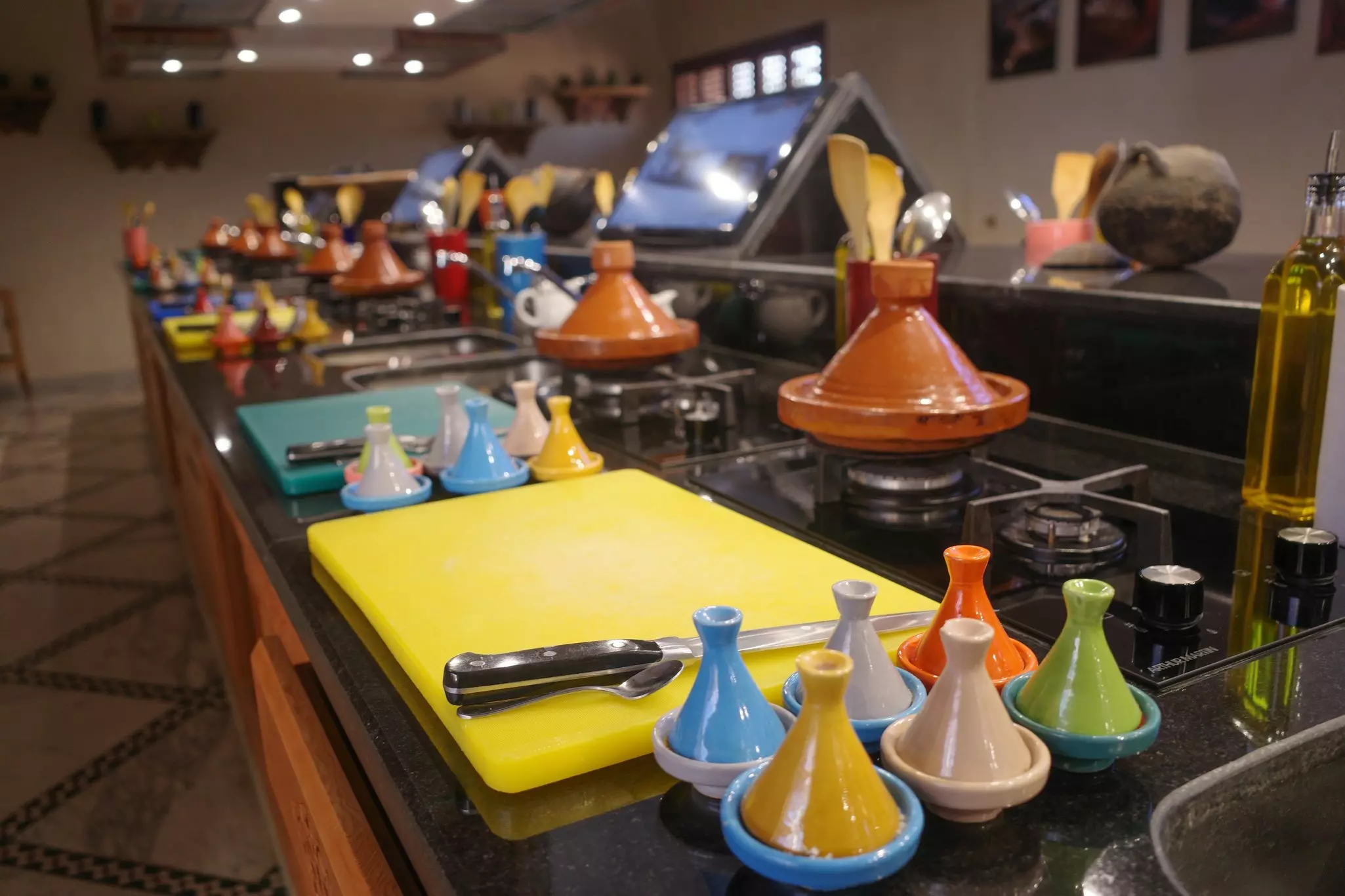 A central counter in a kitchen with multiple cooking spaces each stocked with chopping boards, small pots and a larger tagine on a gas ring.