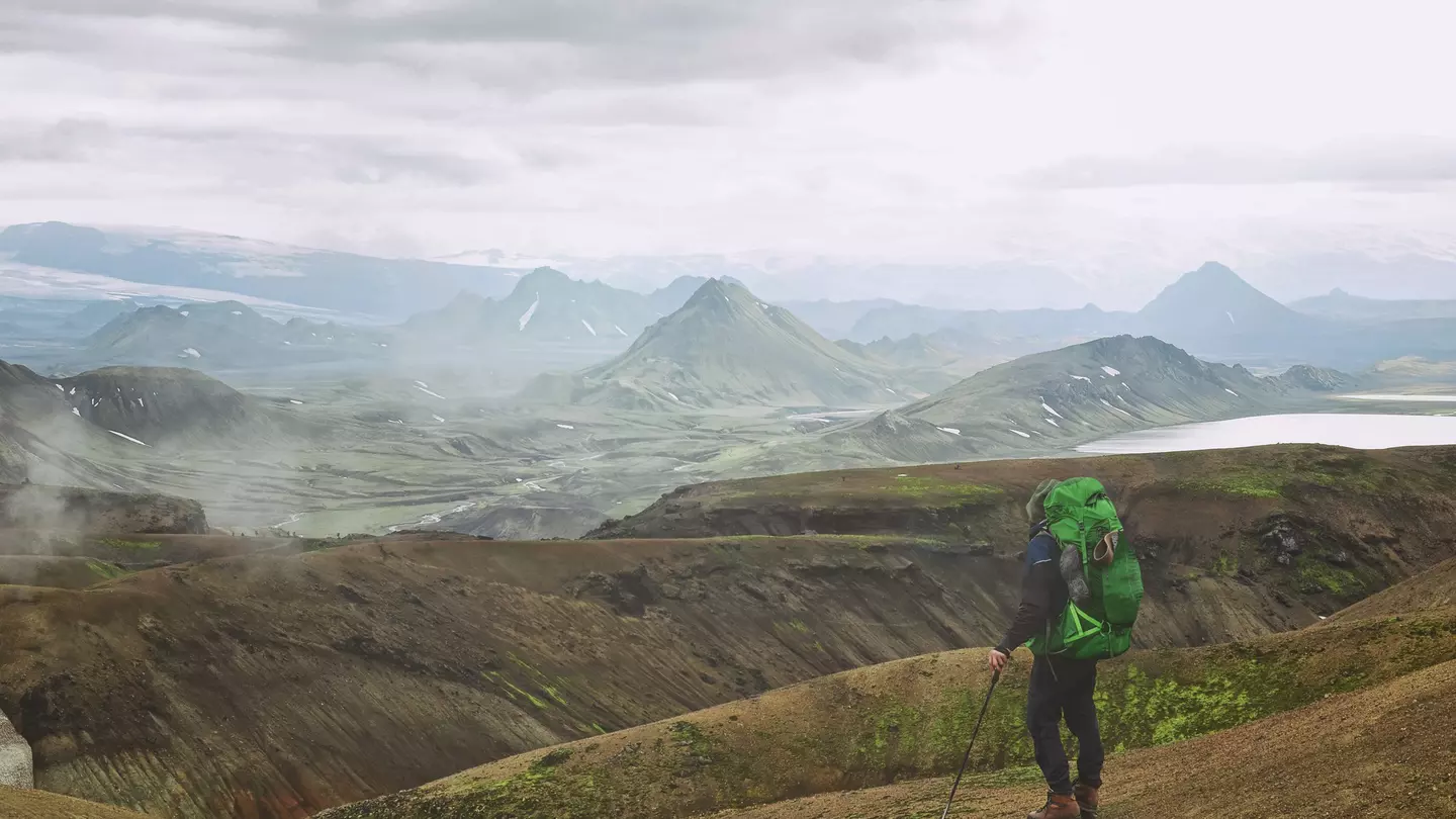 A hiker with a green backpacker looks at mountains across a barren landscape in Iceland.