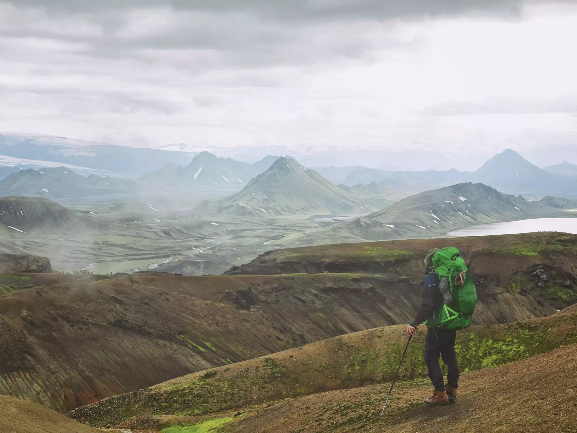 A hiker with a green backpacker looks at mountains across a barren landscape in Iceland.