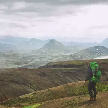 A hiker with a green backpacker looks at mountains across a barren landscape in Iceland.