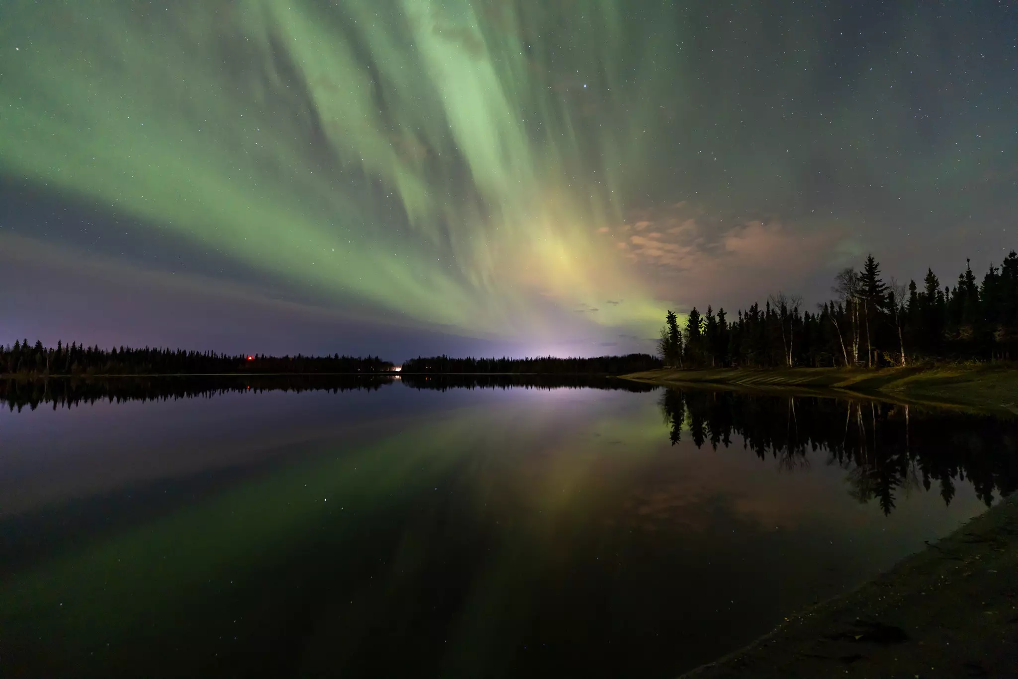Green, purple and yellow lights streak across the night sky, reflected in a lake surrounded by woodland.