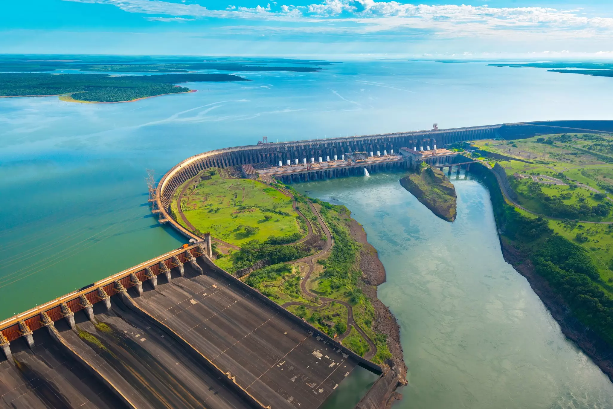 An aerial overshot of a monumental dam, with spillways and hydroelectric turbines visible. A expanse of water is seen behind the dam.