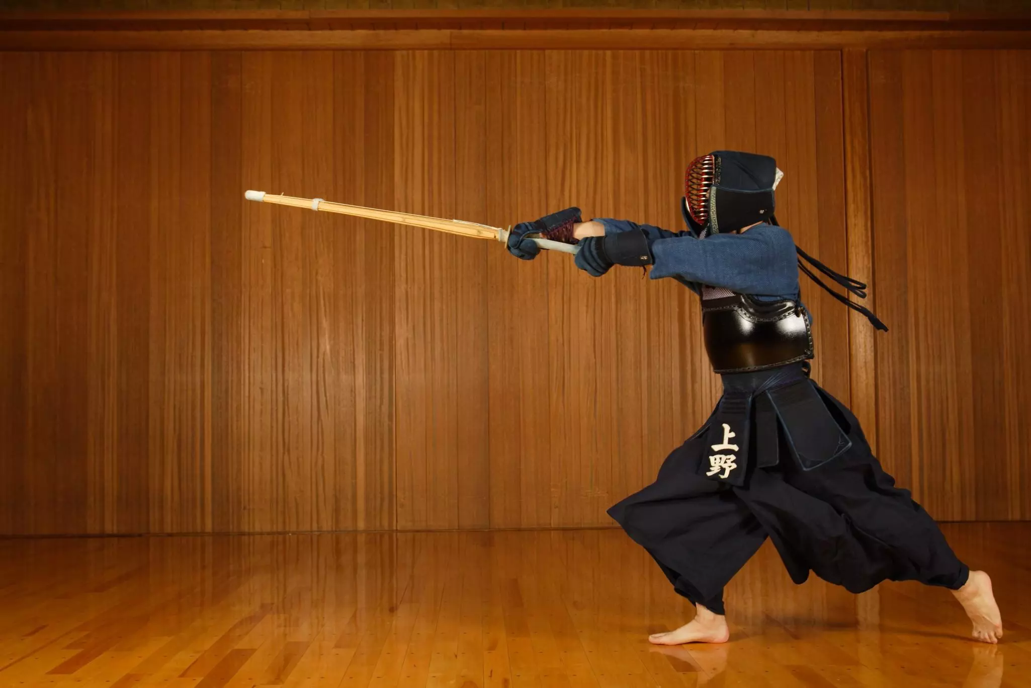 A person dressed in full kendo gear has a fighting stance and holds out the wooden sword