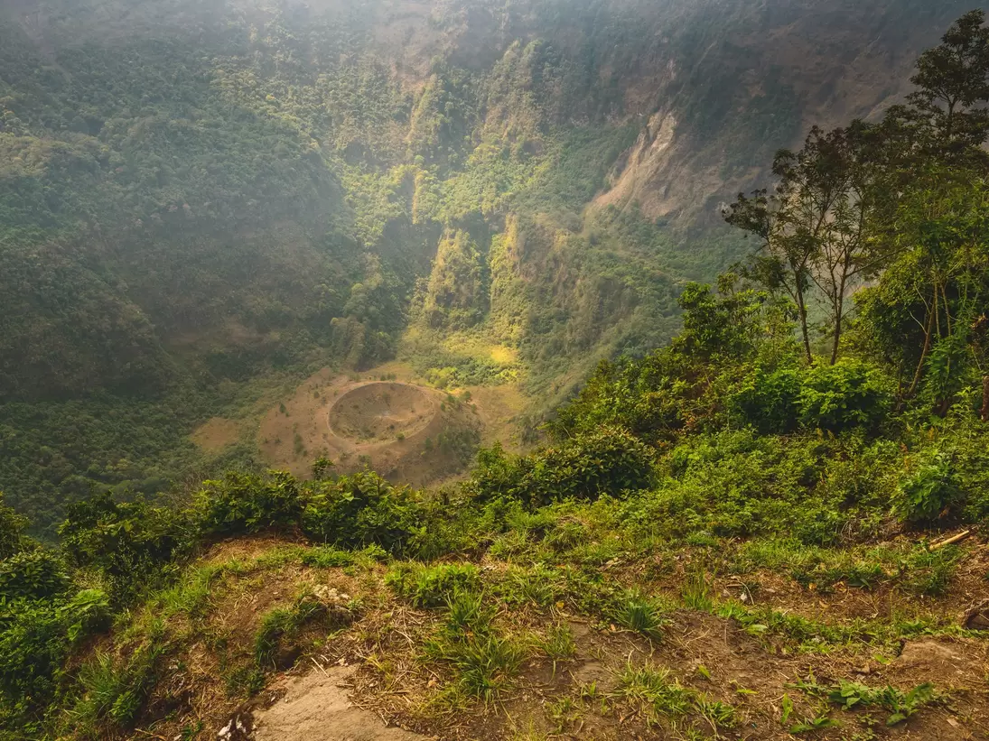 Steep forested crater walls dropping to a bare floor with a small round cone, shrubs on the rim in front