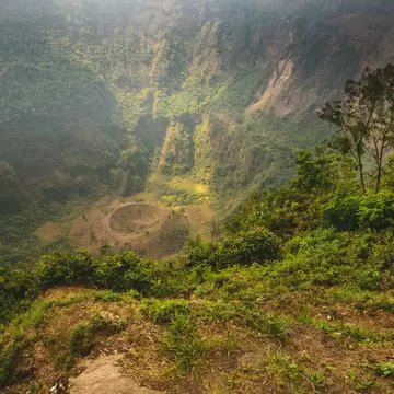 Steep forested crater walls dropping to a bare floor with a small round cone, shrubs on the rim in front