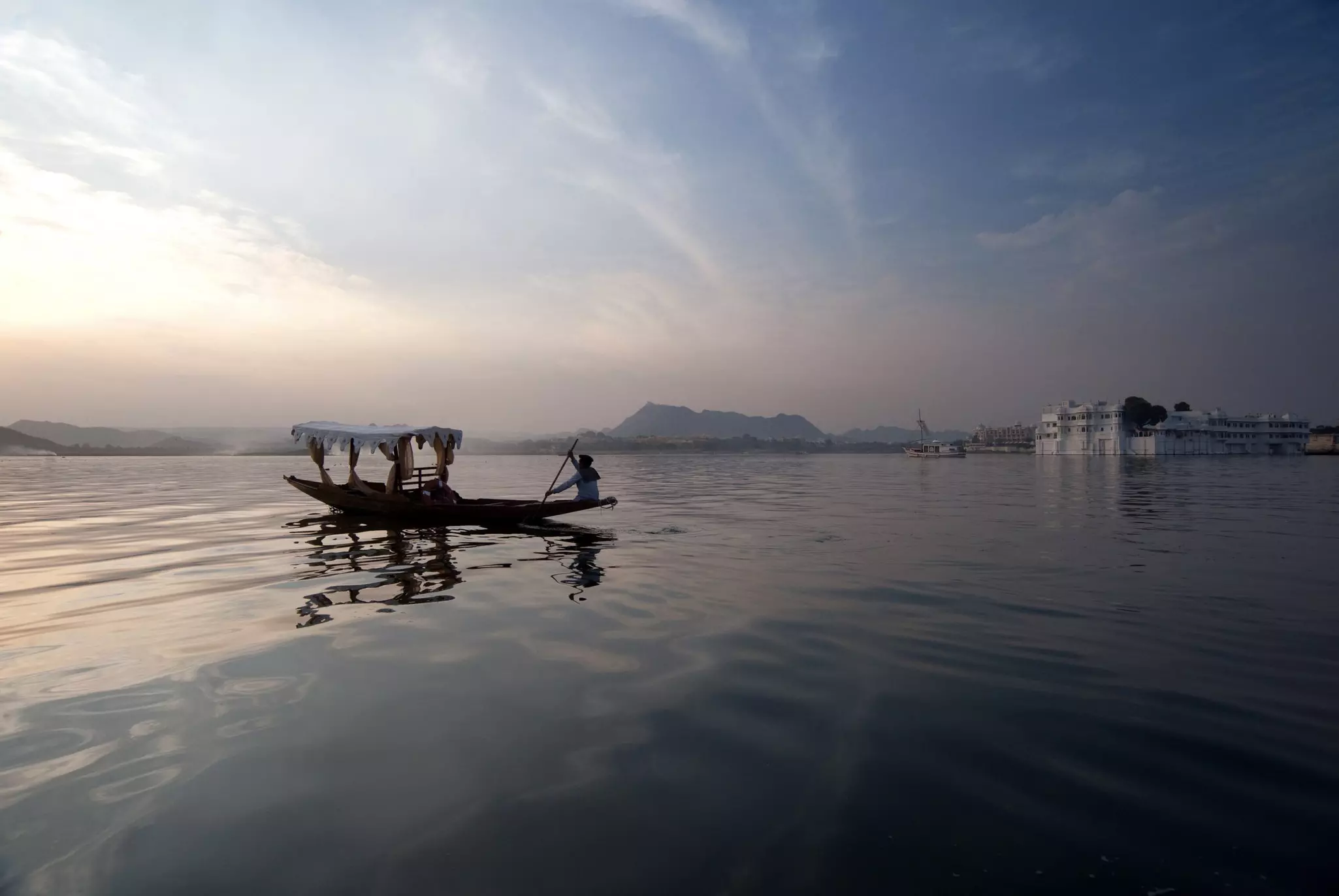 A man paddles a narrow boat on a placid lake. A white palace on an island is visible in the distance.