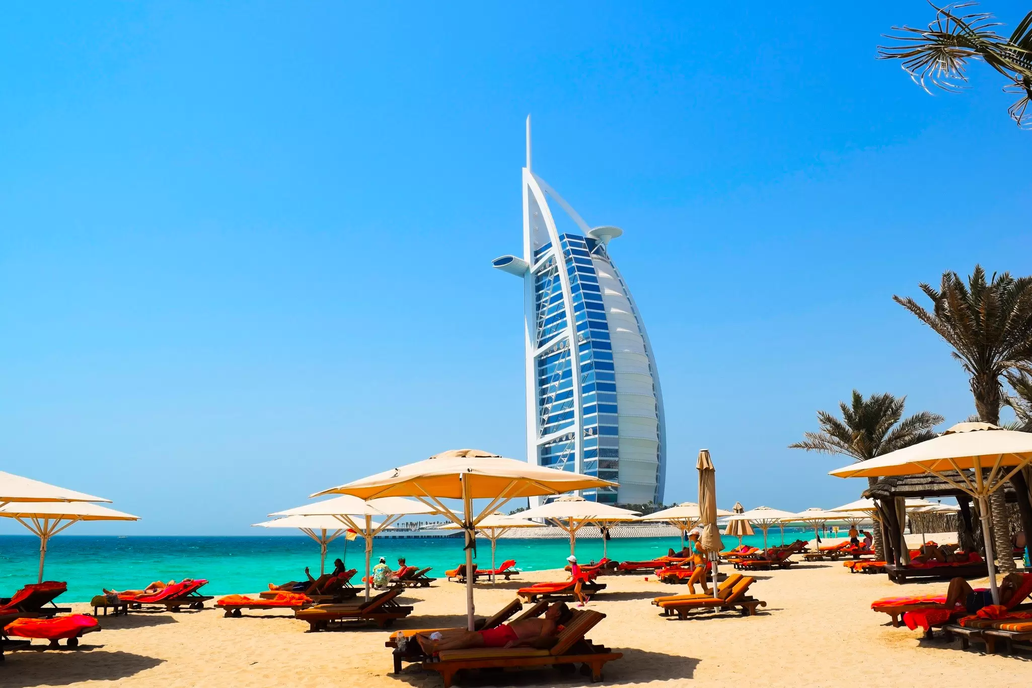 The sail-shaped Burj Al Arab hotel in Dubai seen from a sandy beach lined with sun loungers and white umbrellas
