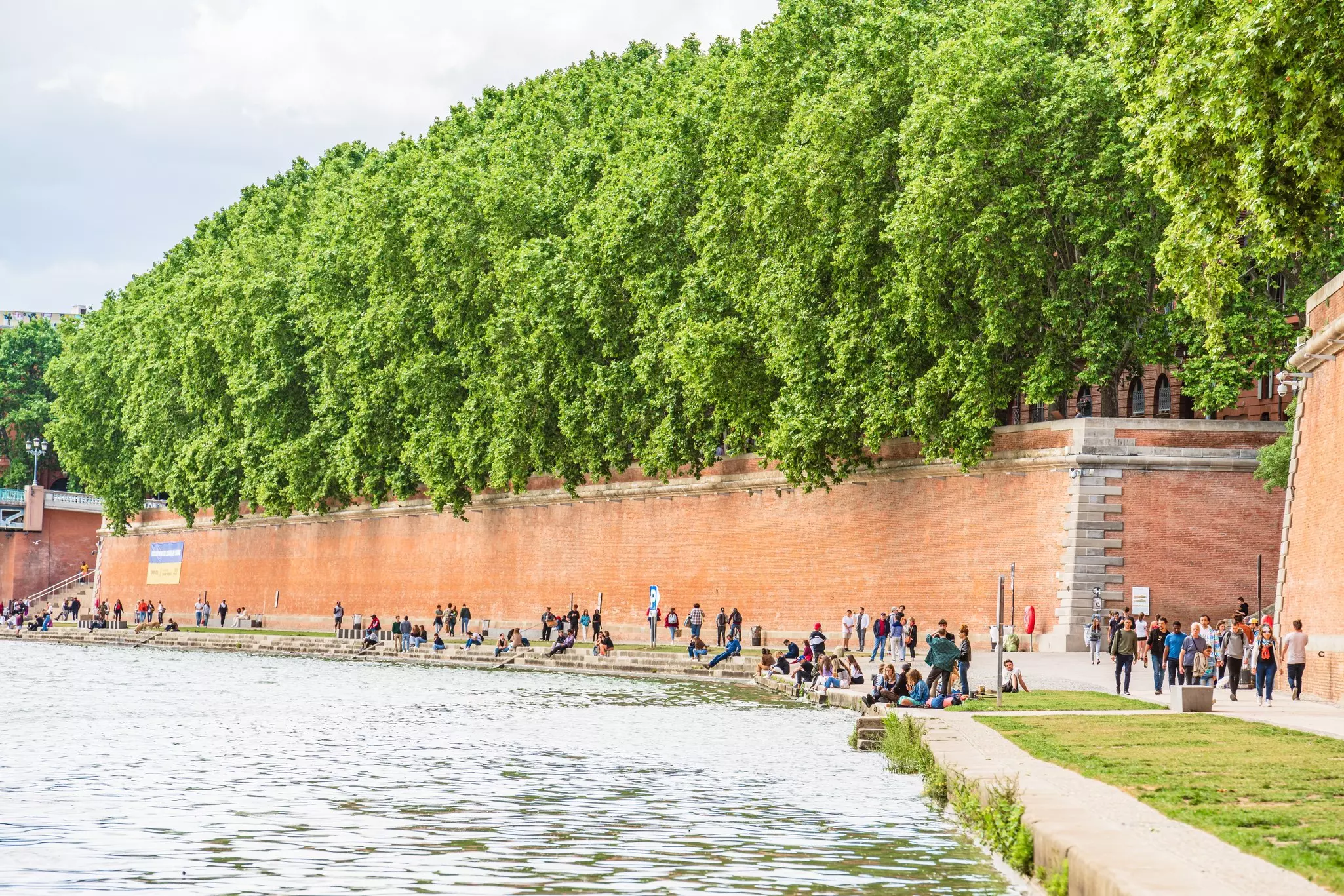 People walking and sitting by a riverbank; there is a tall redbrick wall with leafy green trees above it.