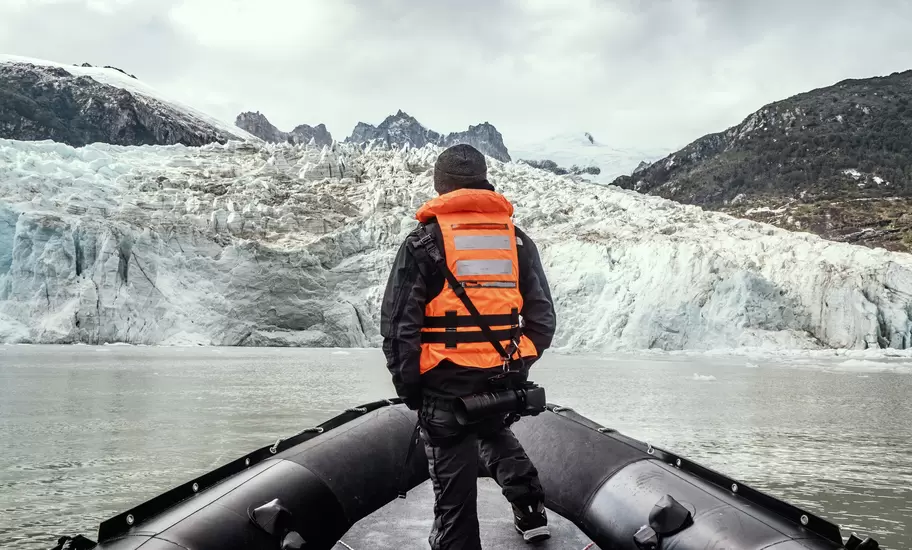 Glacier face meeting grey water in Chilean Patagonia, seen past a person in an orange life vest at the bow of a Zodiac