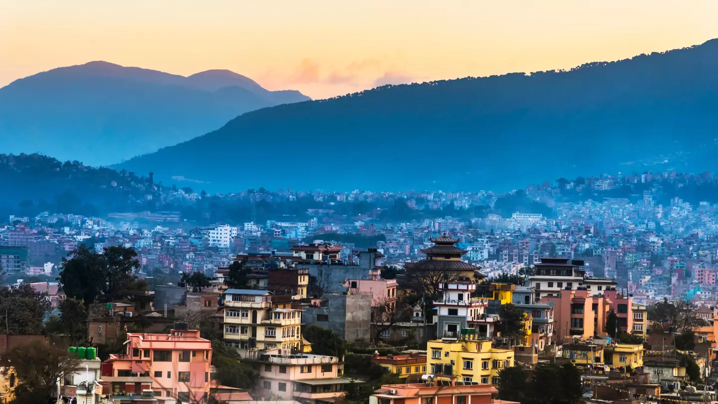 A city with pastel-colored buildings nestled in the foothills of a vast series of mountains.