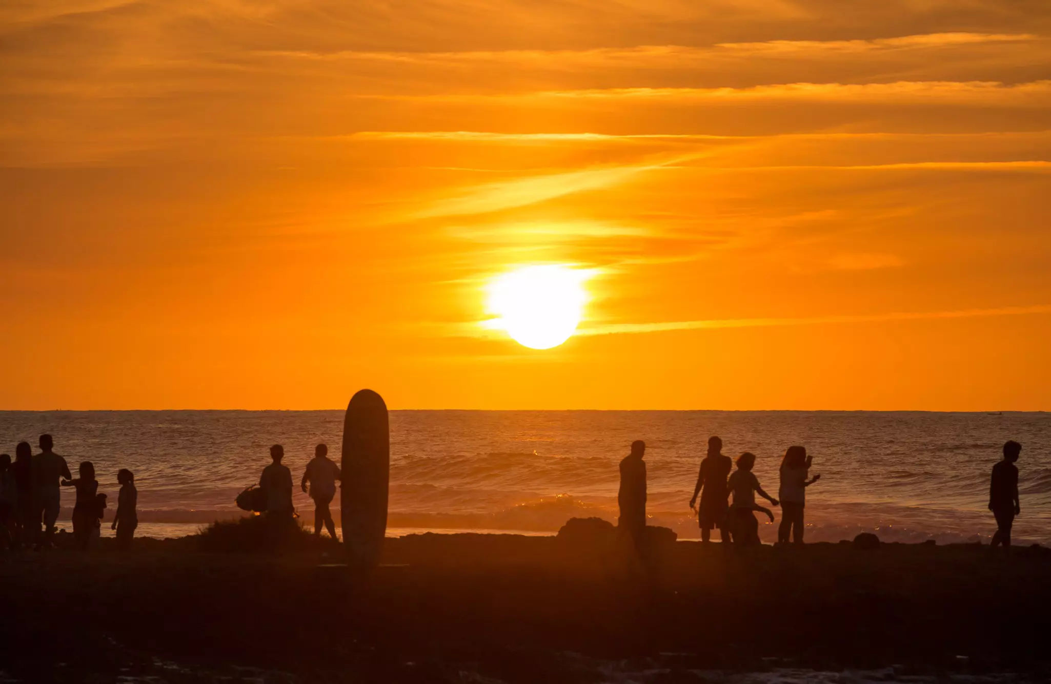 The sun as a fiery orange ball sinking towards the ocean. People in silhouette stand on the beach to watch.