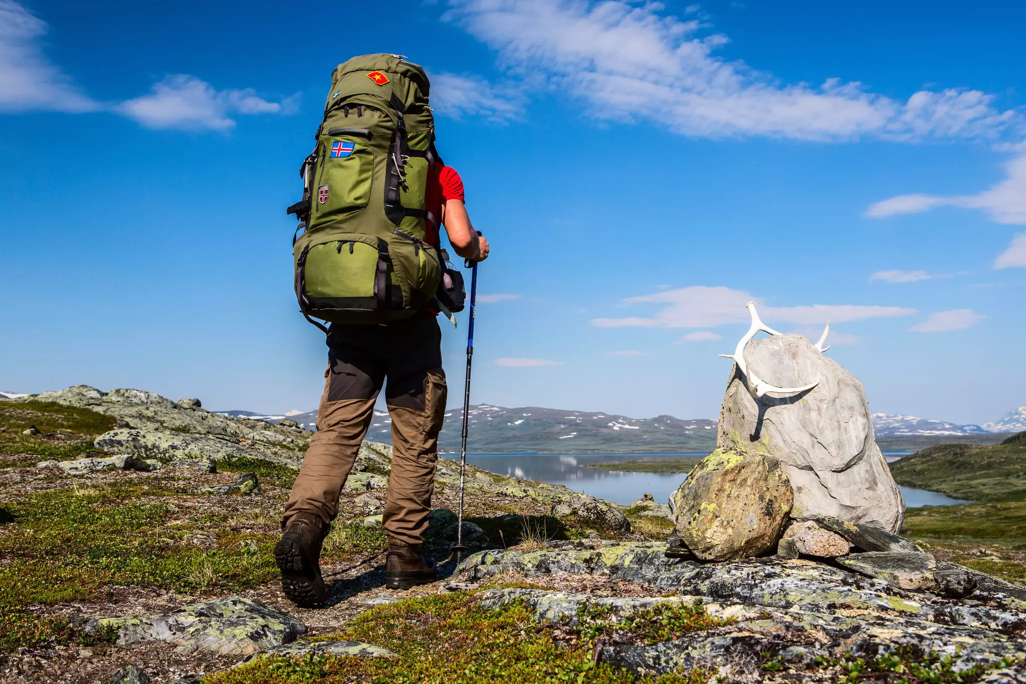 A hiker with a large backpack is seen from behind on the ridge of a hill overlooking a lake. A rock topped with a pair of antlers is to the hiker’s right.