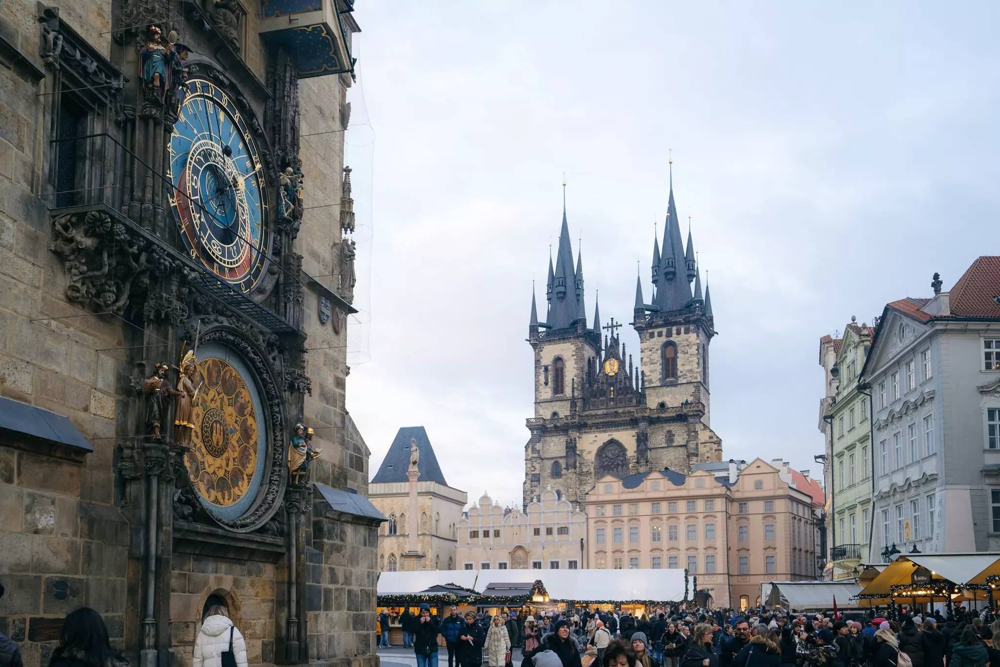An elaborate clock on the side of a building by a square crowded with people; a building with two spires is across the square.