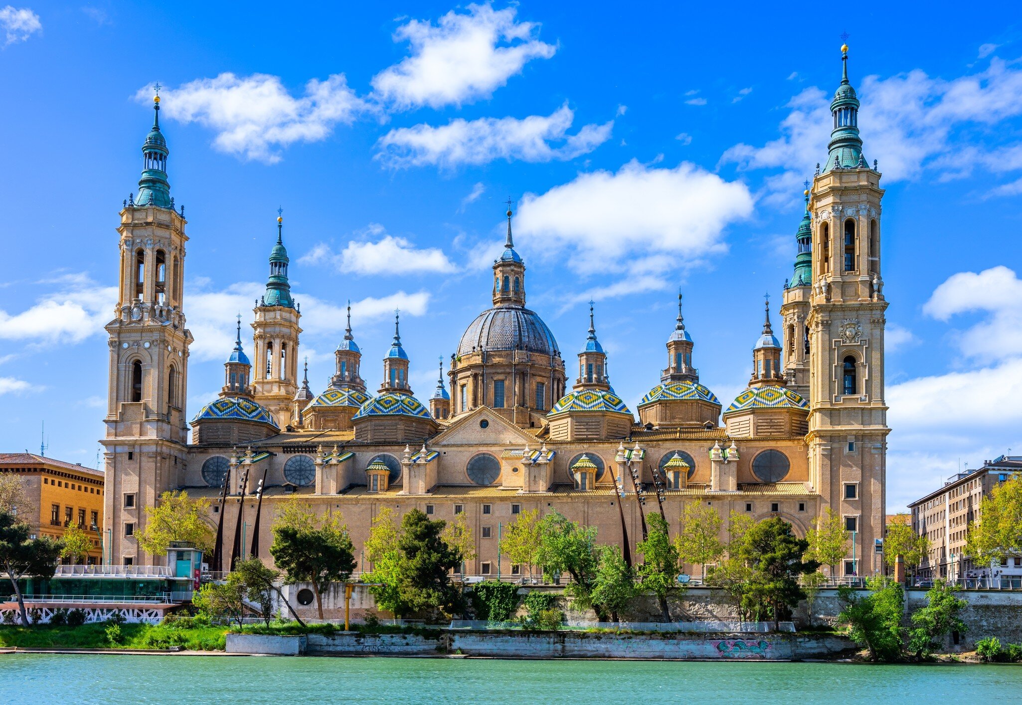 Basílica del Pilar in Zaragoza, Spain: four bell towers and green, yellow and blue tiled domes seen across the river