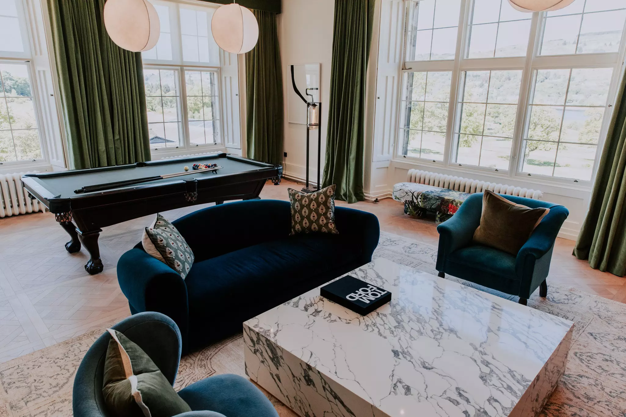 A luxury hotel suite has plush furniture, a pool table, and large windows with velvet drapes.