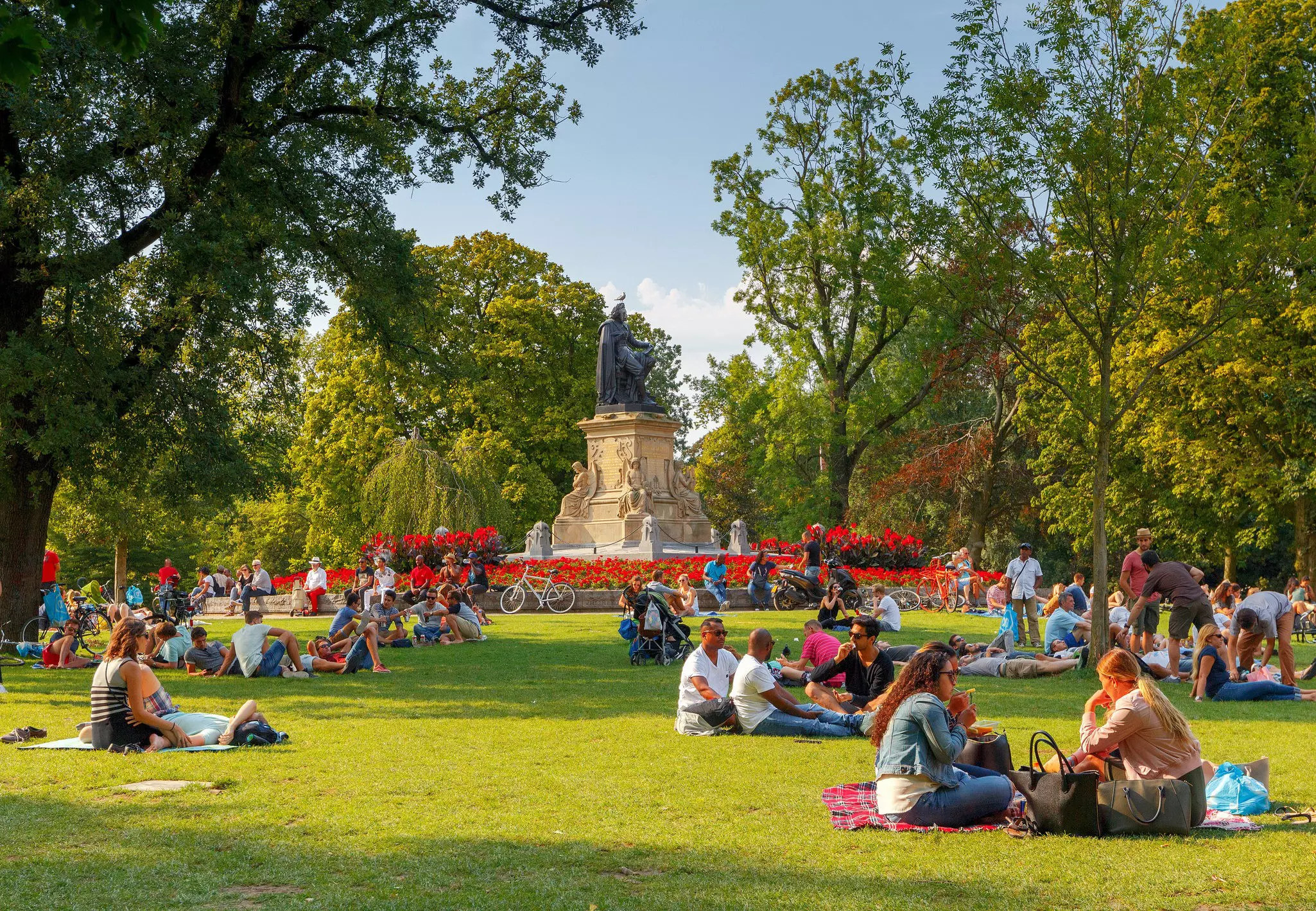 People sat on lawns in a vast park with statues and flowerbeds