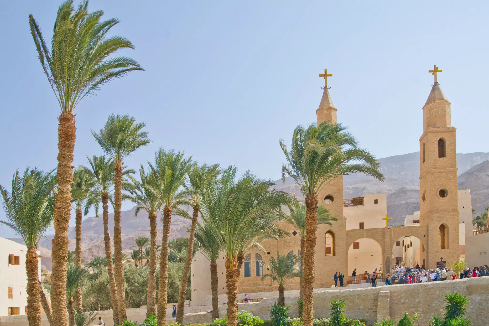 Pilgrims enter a monastery surrounded by palm trees in a hilly desert area.